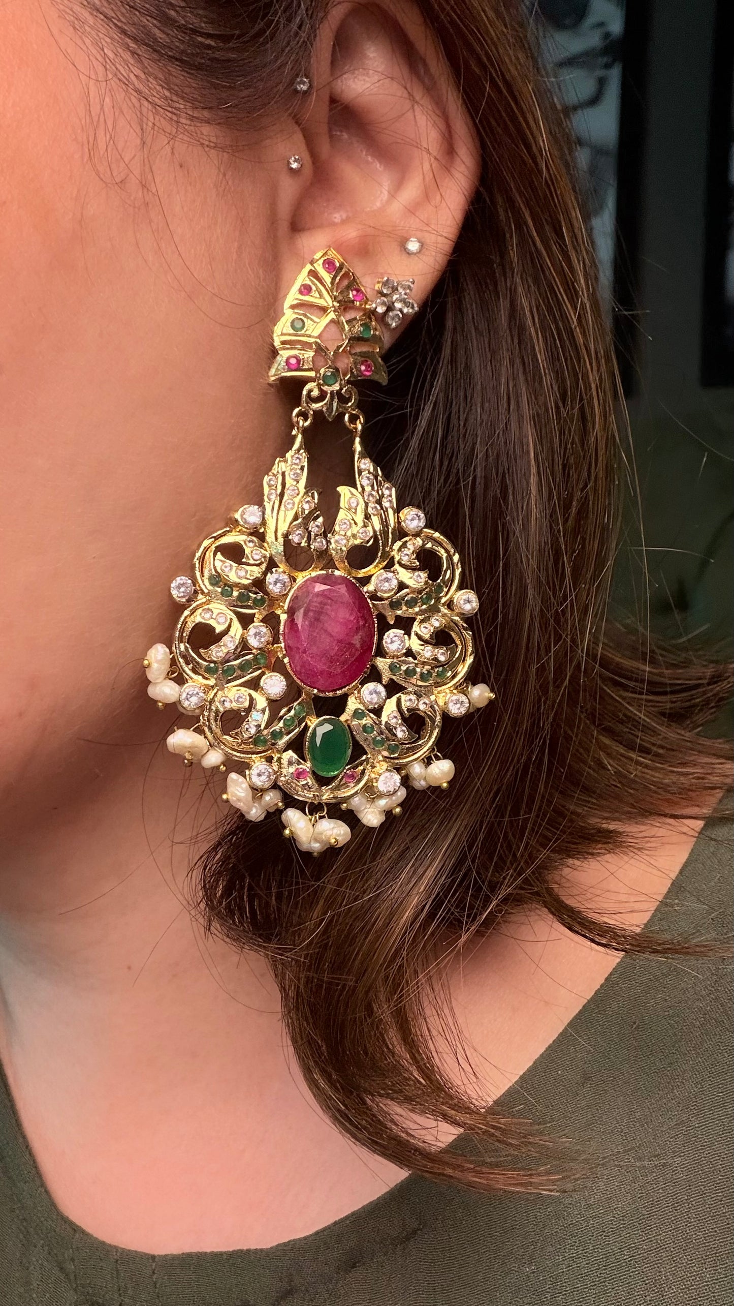 A person wearing an ornate gold-toned statement earring with a pink oval center stone, green accent stones, and small pearl-like beads.