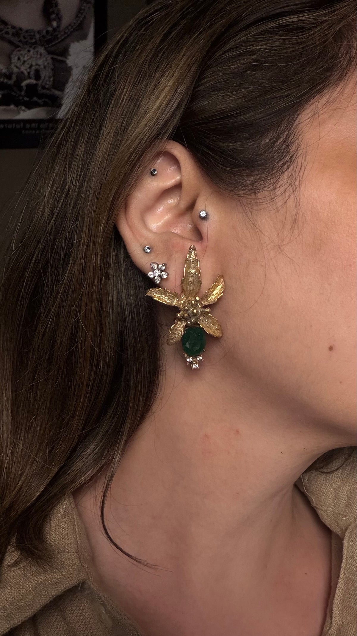 A close-up of a person's ear wearing a gold flower earring with a green gemstone and small clear stones, along with other ear piercings.