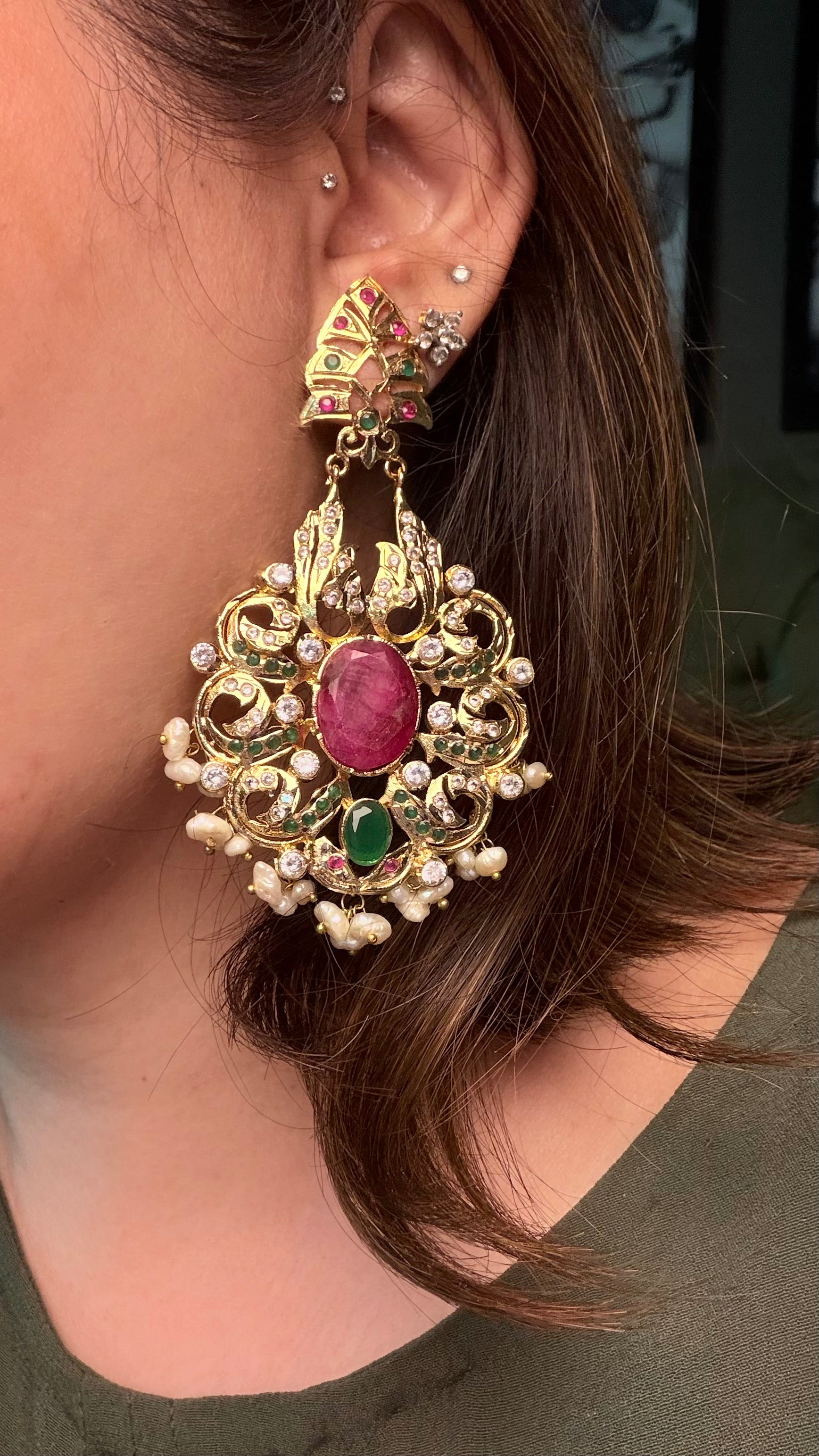 A person wearing an ornate gold-toned statement earring with a pink oval center stone, green accent stones, and small pearl-like beads.